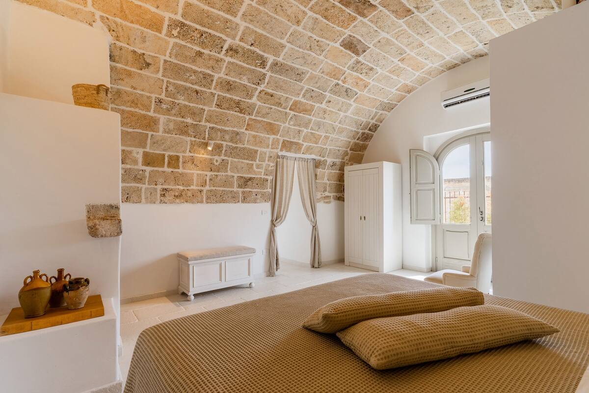 A bedroom with a stone ceiling, a bed with beige bedding, a wooden shelf with pottery, a white wardrobe, and a window with curtains.