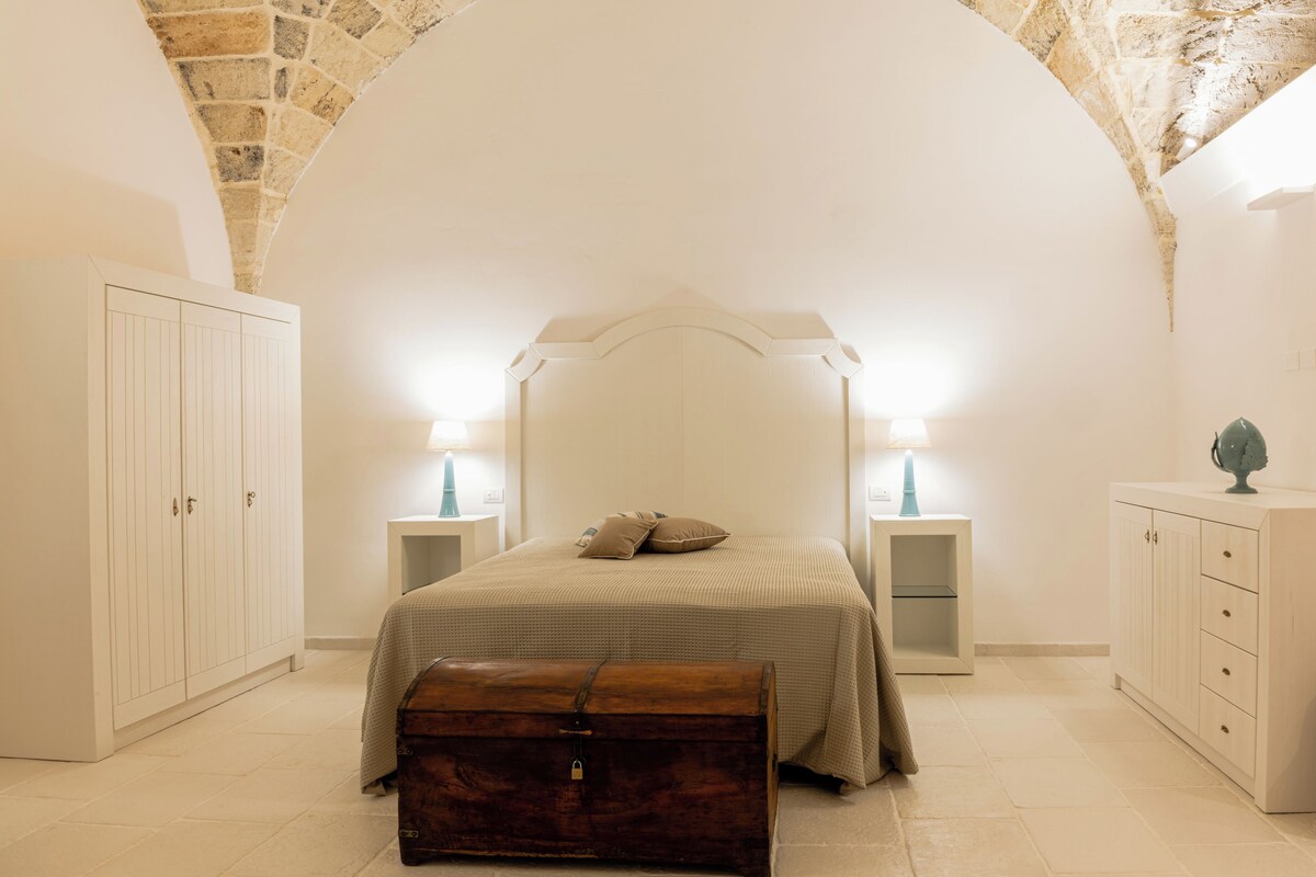 Quarto com teto em arco de pedra, cama com cabeceira, mesas de cabeceira com abajures, guarda-roupa e cômoda.