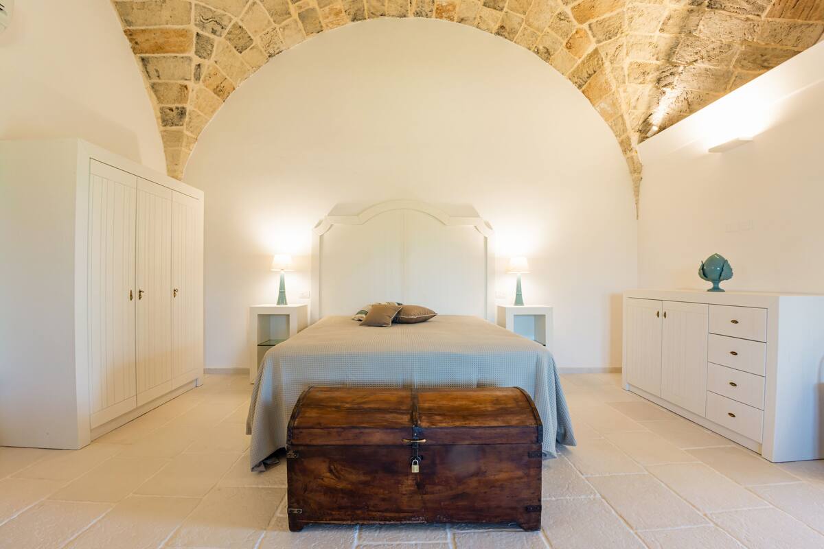 A bedroom with a stone ceiling, a bed with a white headboard, a wooden chest, and white cabinets.