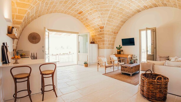A spacious living area with a stone ceiling, a kitchen counter with bar stools, a seating area with a sofa and chairs, and a television mounted on the wall. - Masseria Macchiaforte (Gallipoli)