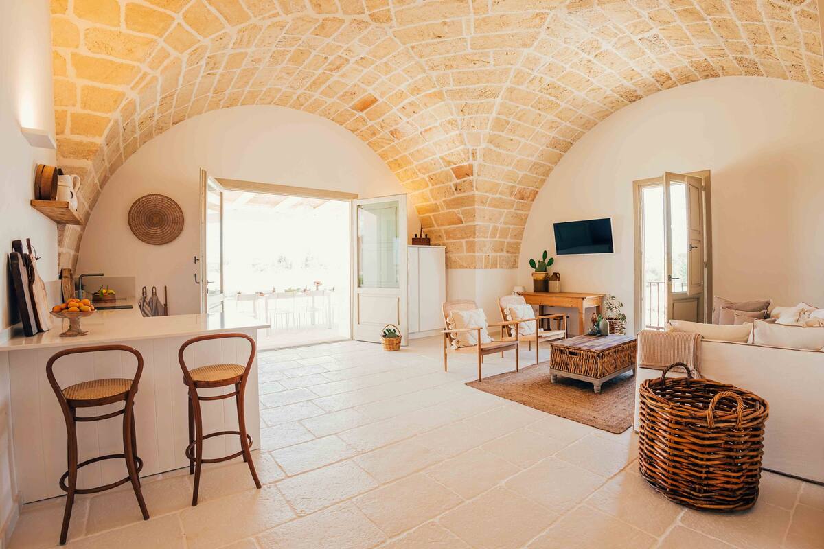 A spacious living area with a stone ceiling, a kitchen counter with bar stools, a seating area with a sofa and chairs, and a television mounted on the wall.