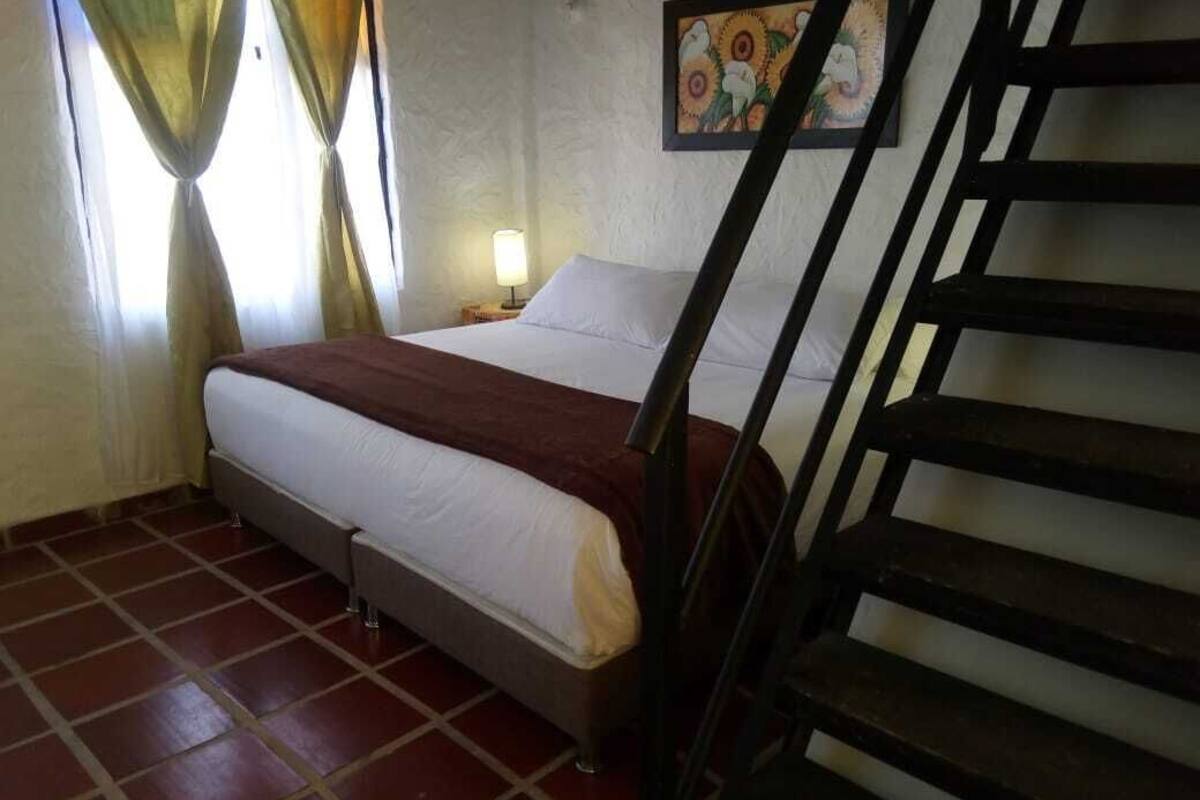 Un dormitorio con cama, mesita de noche, lámpara, un cuadro en la pared y una escalera.