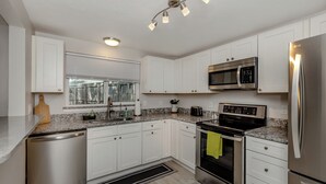 A modern kitchen with white cabinets, stainless steel appliances, a granite countertop, and a window with blinds. - The Lemon Bay Getaway (Venice)