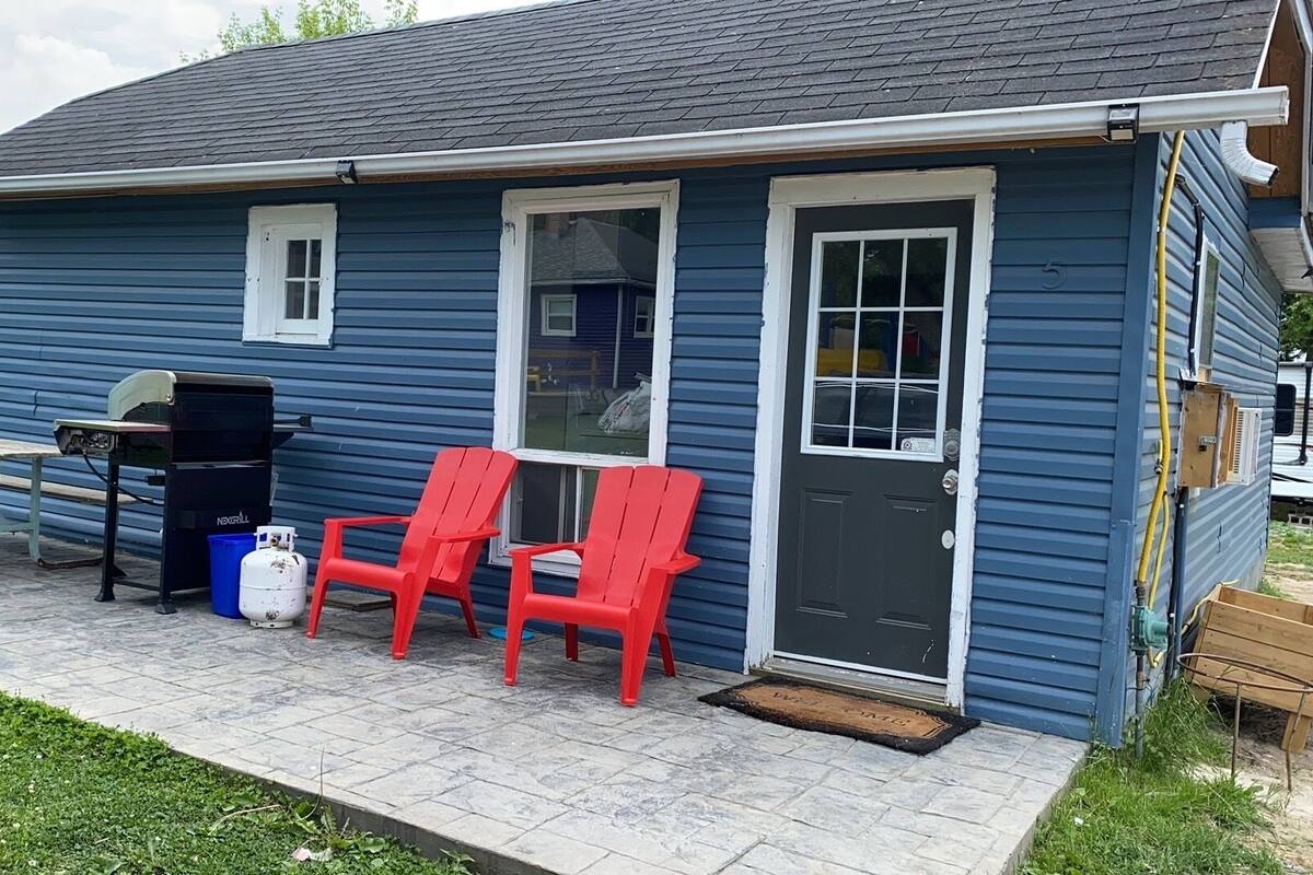 A small blue house with a front porch, a grill, and two red Adirondack chairs.