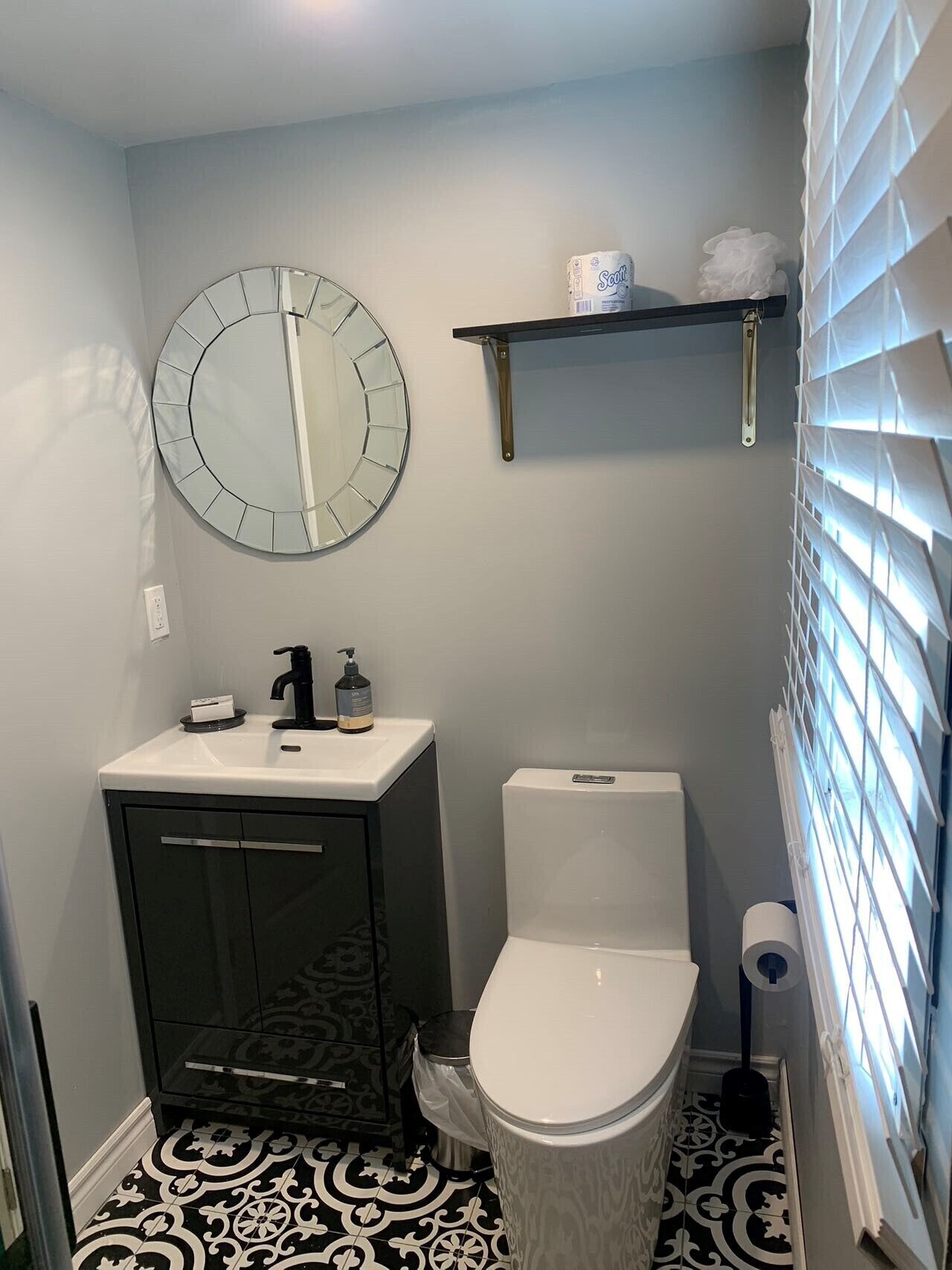 Une salle de bain avec un miroir rond, des toilettes, un lavabo avec un distributeur de savon et un porte-rouleau de papier toilette.