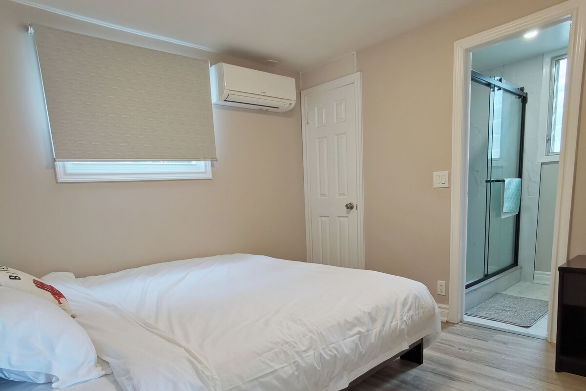 A bedroom with a bed, a window with blinds, an air conditioning unit, and a door leading to a bathroom.