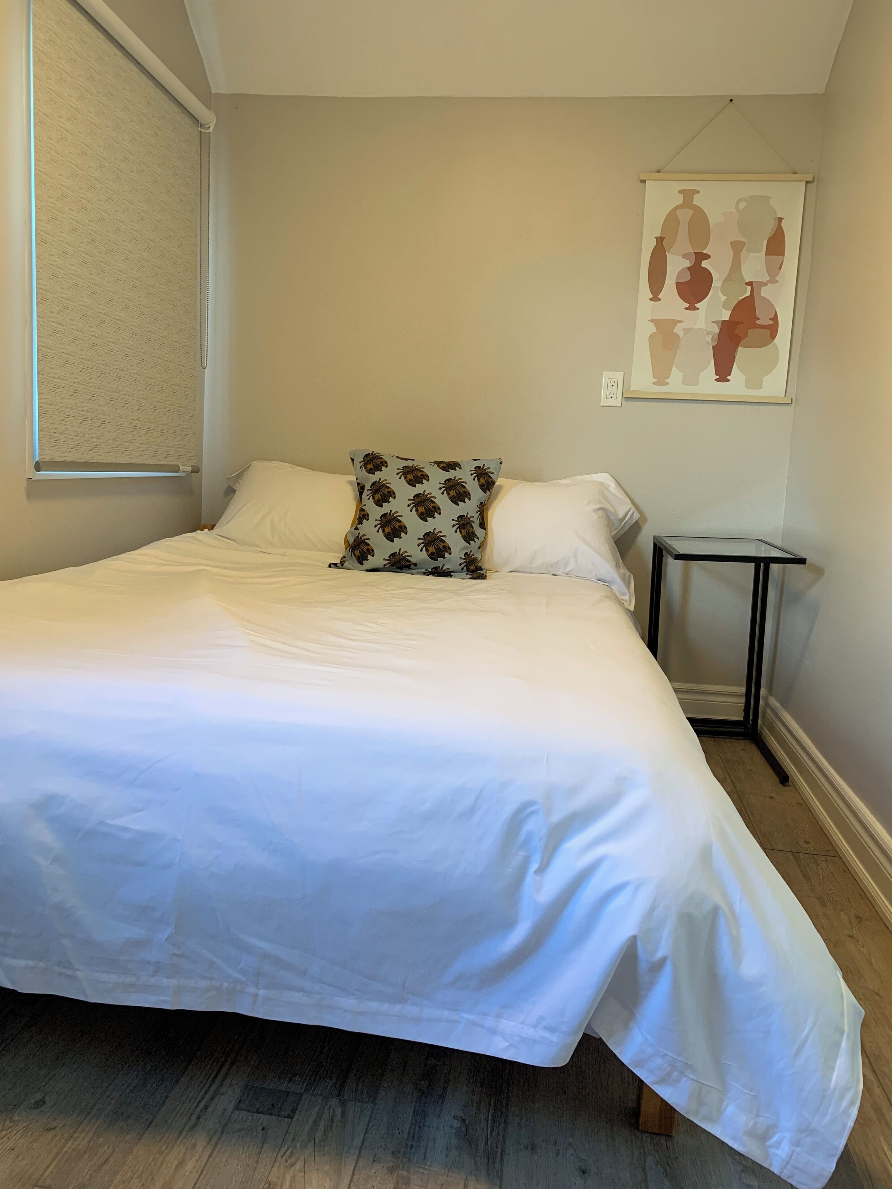 A neatly made bed with white linens, a pillow with a palm leaf pattern, a small bedside table, and a wall-mounted artwork of vases.