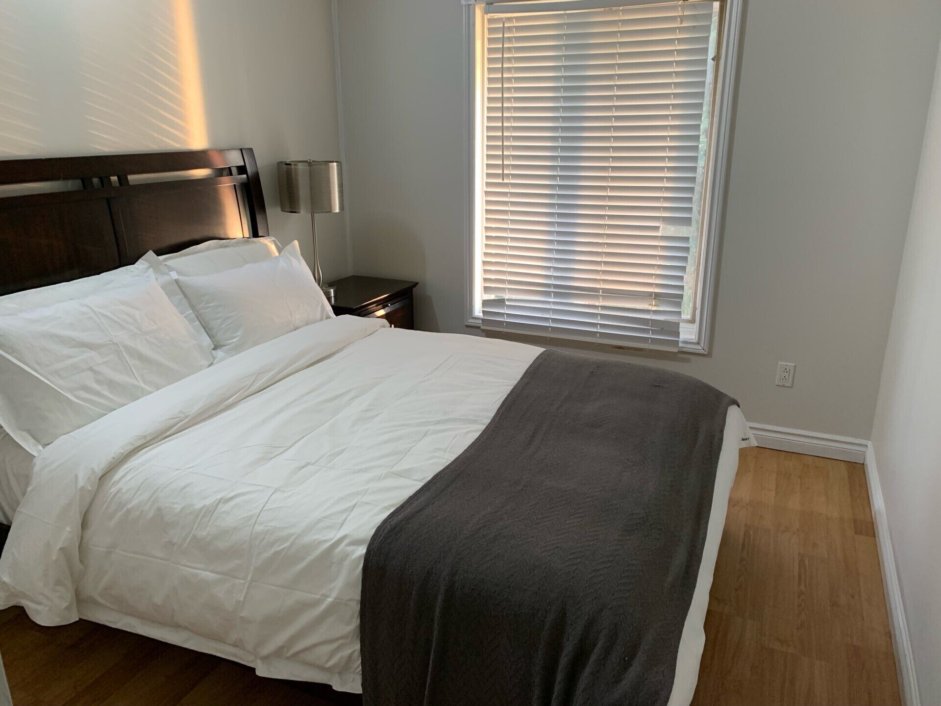 Una cama bien hecha con ropa blanca y una manta gris, una mesita de noche con lámpara, y una ventana con persianas.