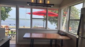 Eine Küche mit Holztisch und Bank, großen Fenstern mit Seeblick und roten Sonnenschirmen, sowie einer Deckenleuchte.