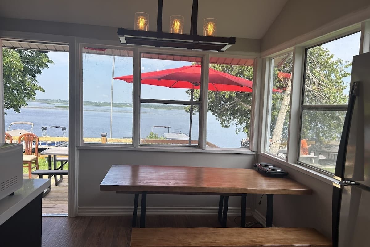 A kitchen with a wooden table and bench, large windows with a view of a lake and red umbrellas, and a ceiling light fixture.