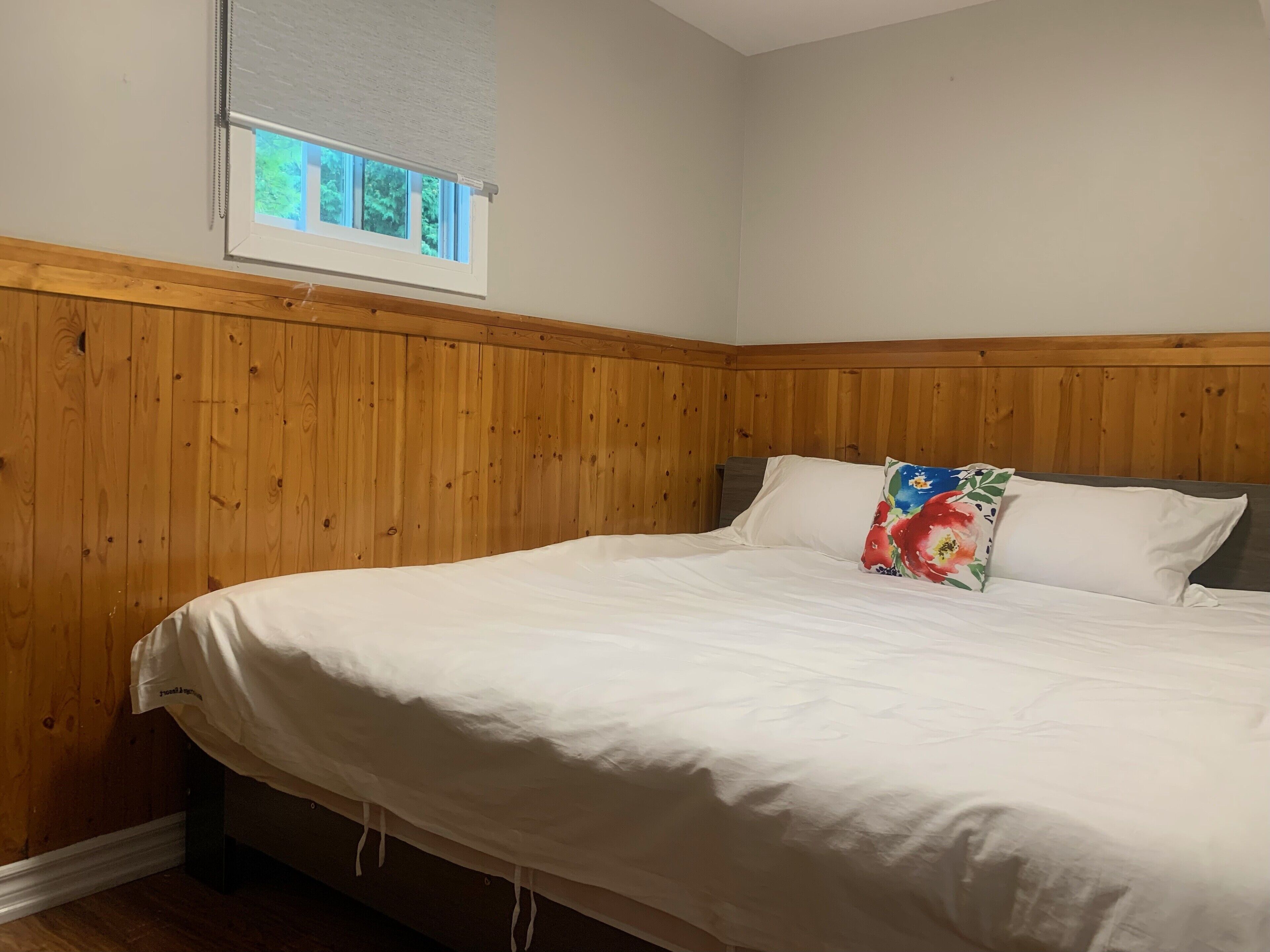 Ein Schlafzimmer mit einer holzgetäfelten Wand, einem Fenster mit Jalousien, einem Bett mit weißer Bettwäsche und einem blumigen Kopfkissen.