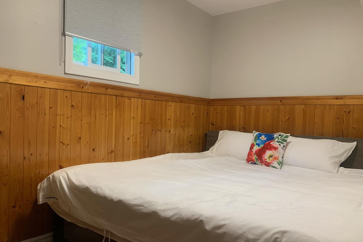 Ein Schlafzimmer mit einer holzgetäfelten Wand, einem Fenster mit Jalousien, einem Bett mit weißer Bettwäsche und einem blumigen Kopfkissen.