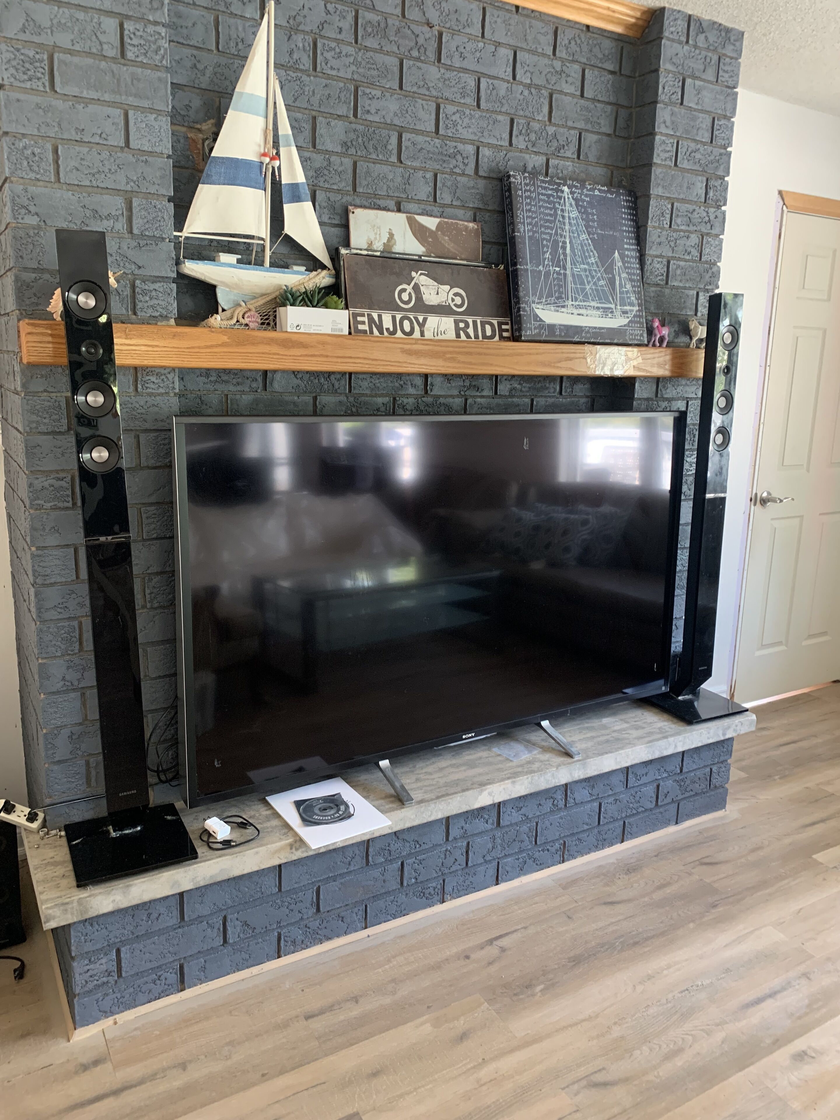 A modern kitchen with a large black flat-screen TV mounted on a brick wall, a wooden shelf above the TV, and a built-in oven below.