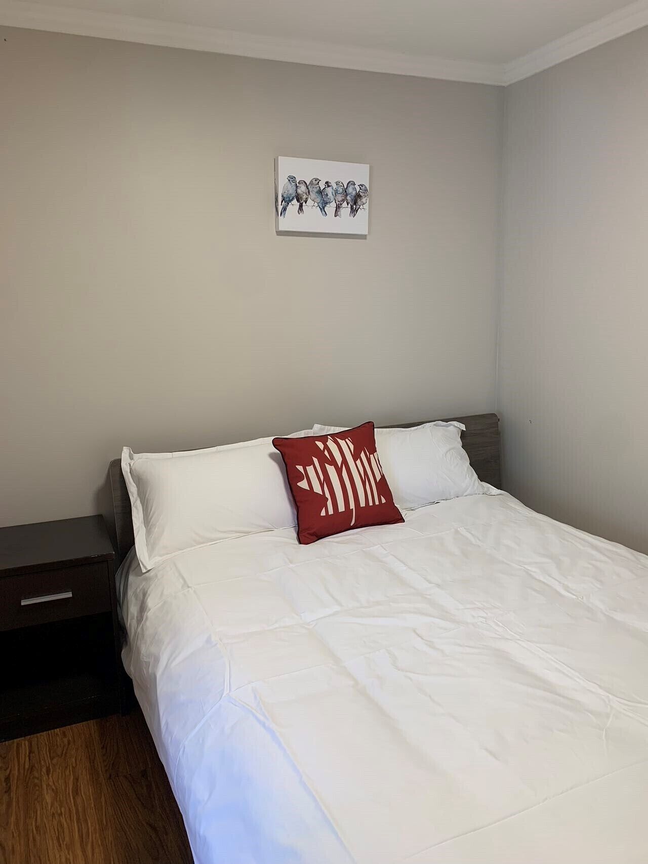 Una cama bien hecha con ropa blanca y una almohada roja, una mesita de noche pequeña y un cuadro con pájaros en la pared.