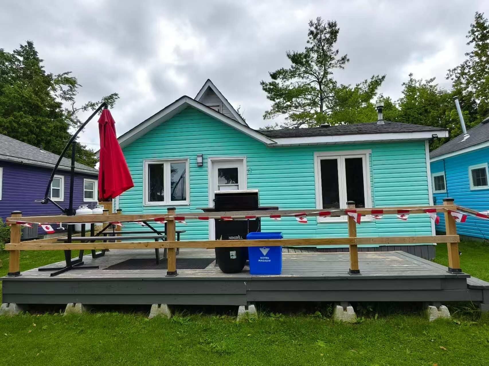 A turquoise house with a deck, a red umbrella, and a blue trash bin labeled "Notre Magazine".