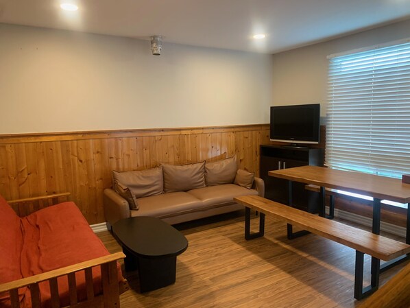 A cozy living area with a wooden panel wall, a sofa, a television, and a dining table with benches. - Myriad Fins Cottage and Marina (Kawartha Lakes)