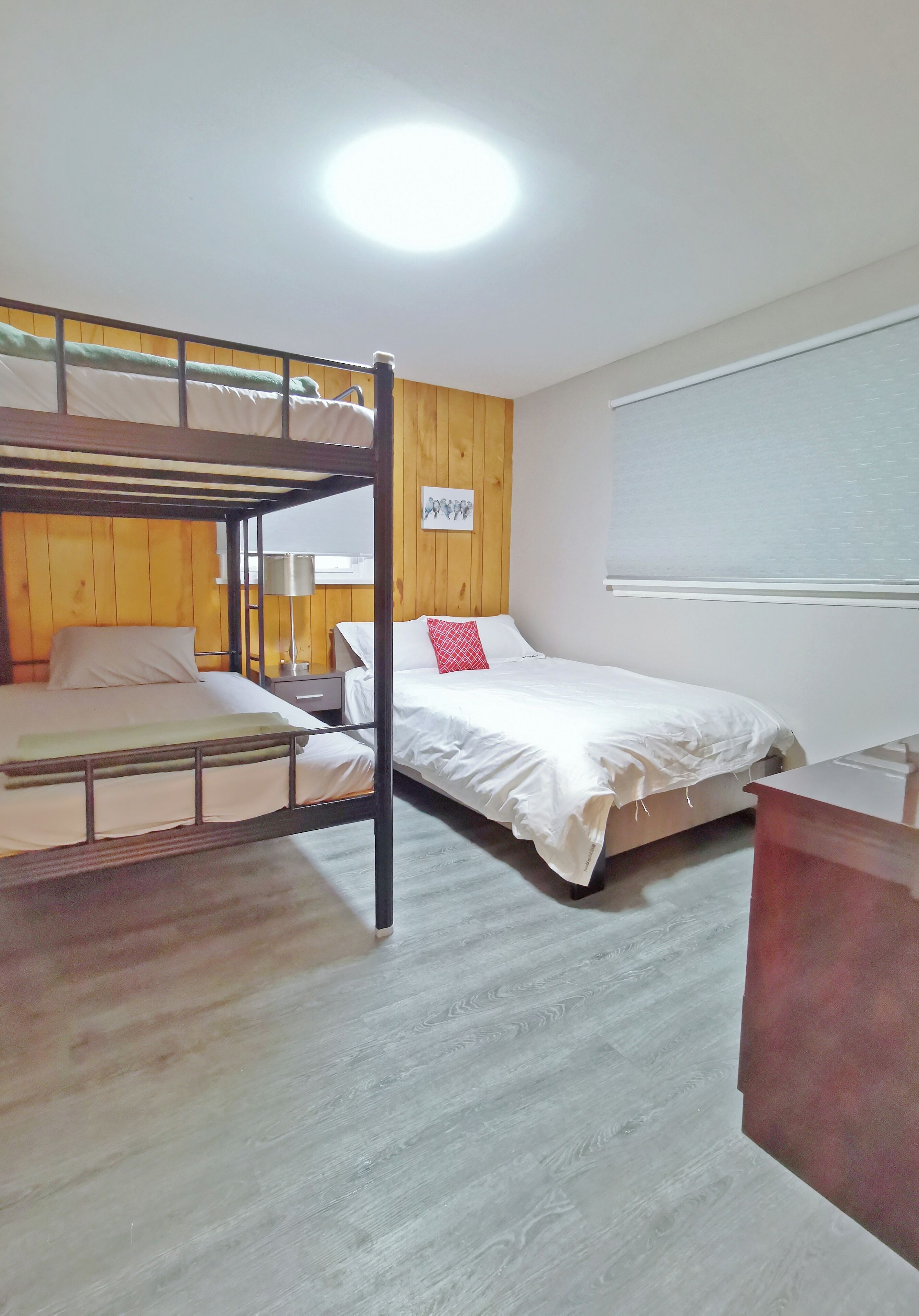 A room with bunk beds, a wooden wall, a window with blinds, and a desk.