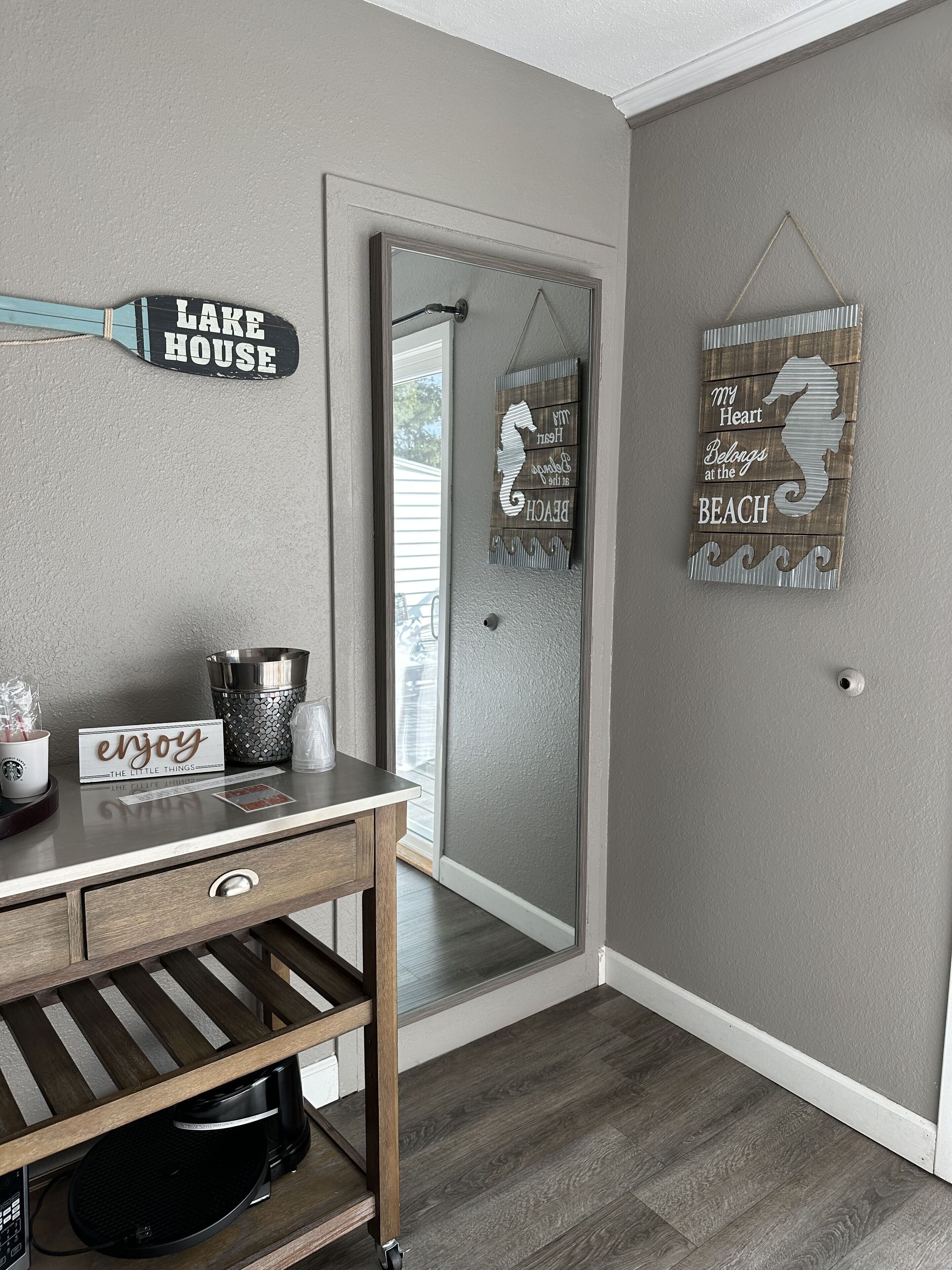 A hallway with a wooden desk, a sign reading "enjoy the little things," and a mirror reflecting a door.
