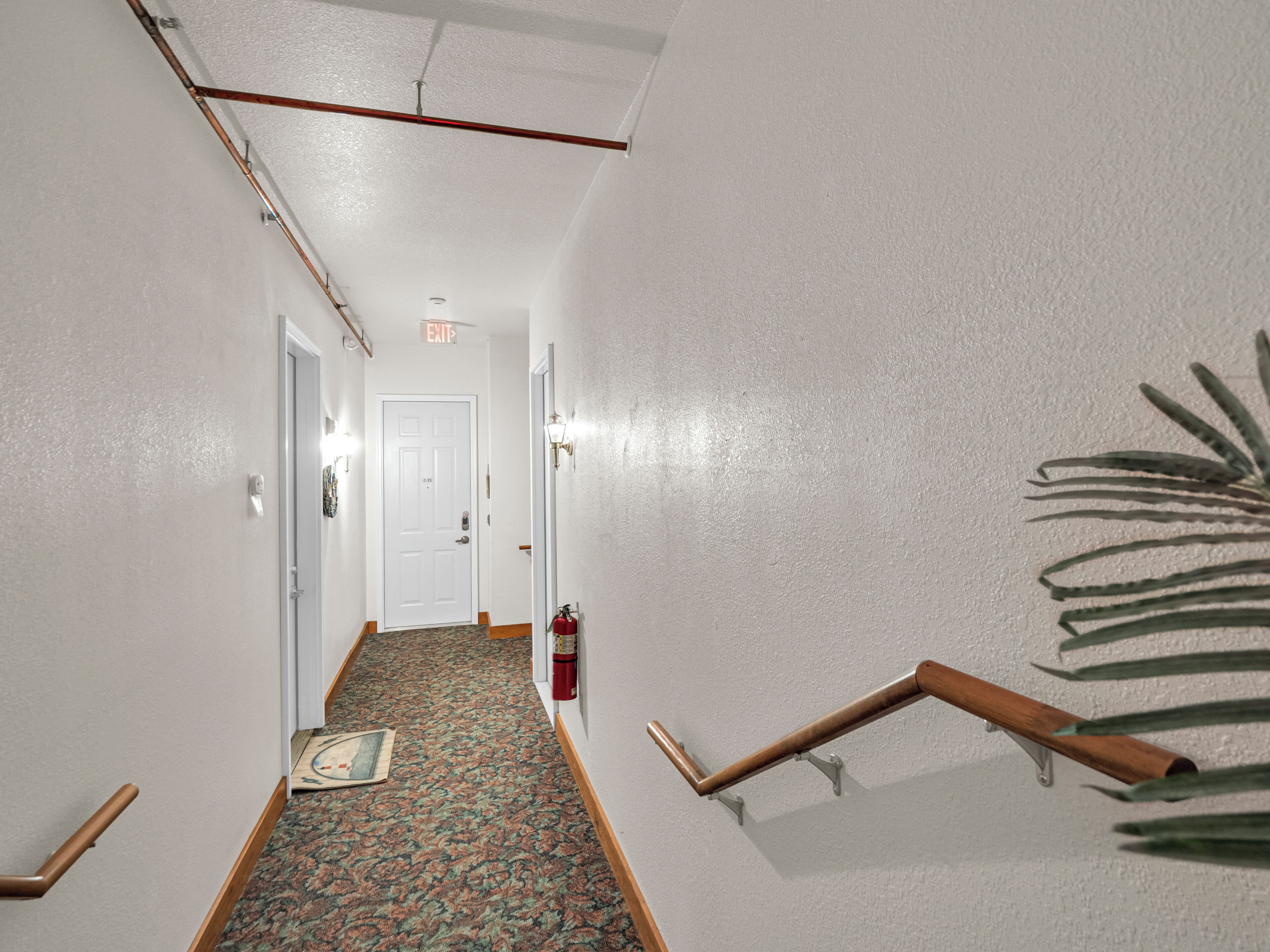A hallway with a patterned carpet, white walls, a white door at the end, and a fire extinguisher mounted on the wall.