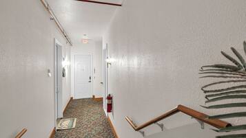 A hallway with a patterned carpet, white walls, a white door at the end, and a fire extinguisher mounted on the wall.
