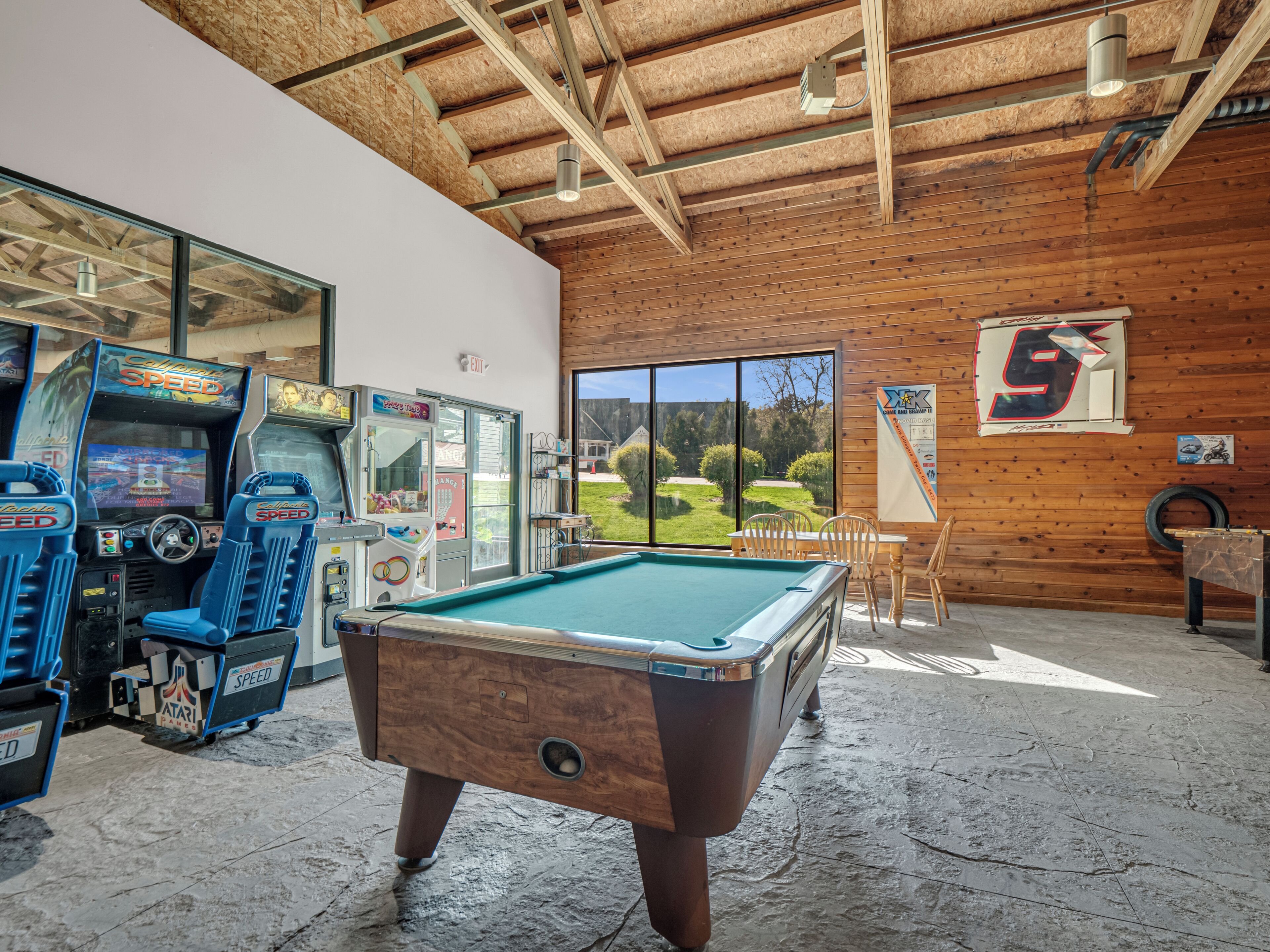 A pool table in a room with wooden walls and ceiling, arcade games, and a view of a golf course.
