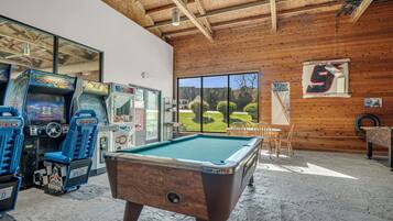 A pool table in a room with wooden walls and ceiling, arcade games, and a view of a golf course.