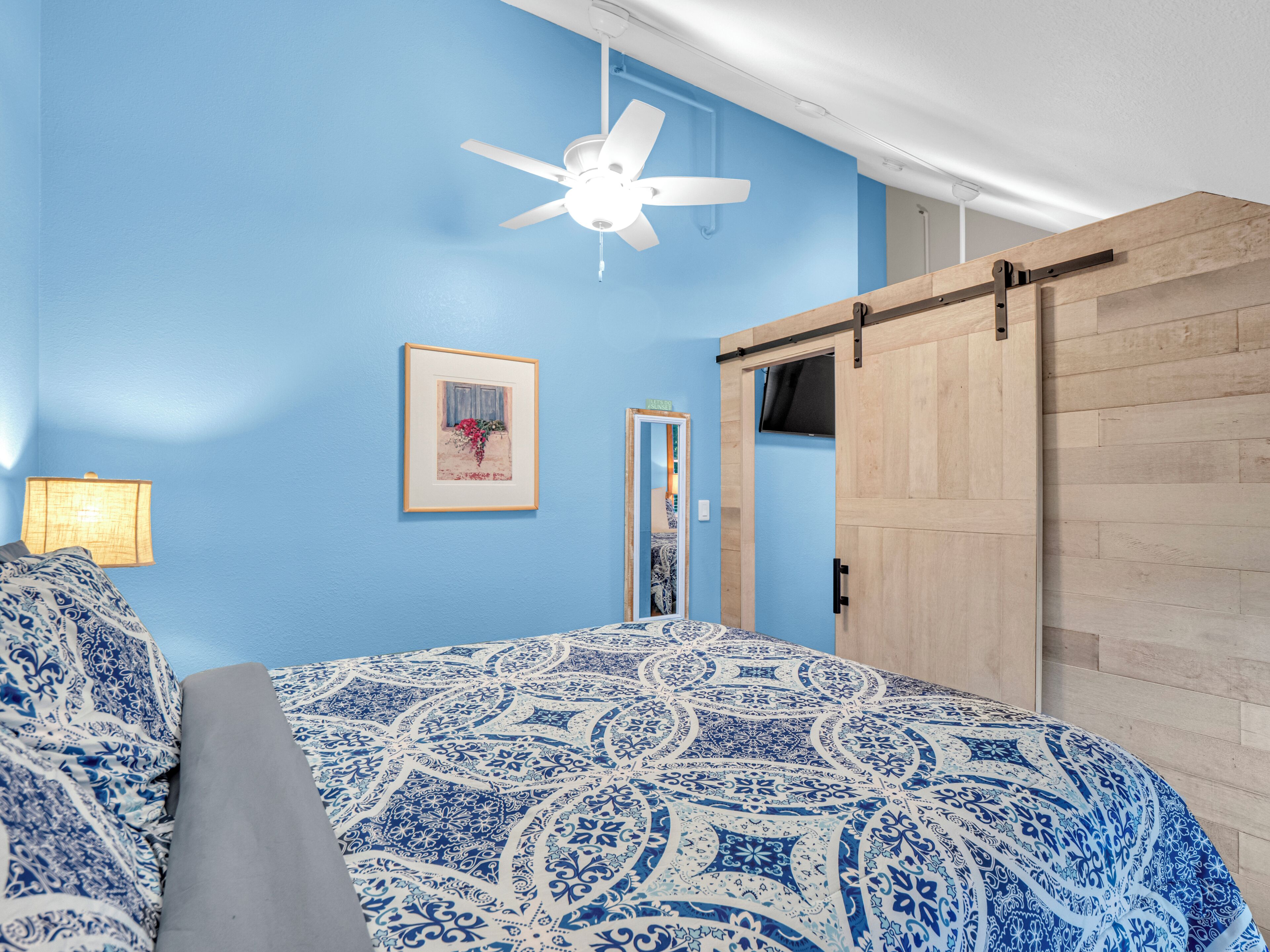 Un dormitorio con pared azul, ventilador de techo, cama con colcha estampada, un cuadro enmarcado y una puerta corredera.