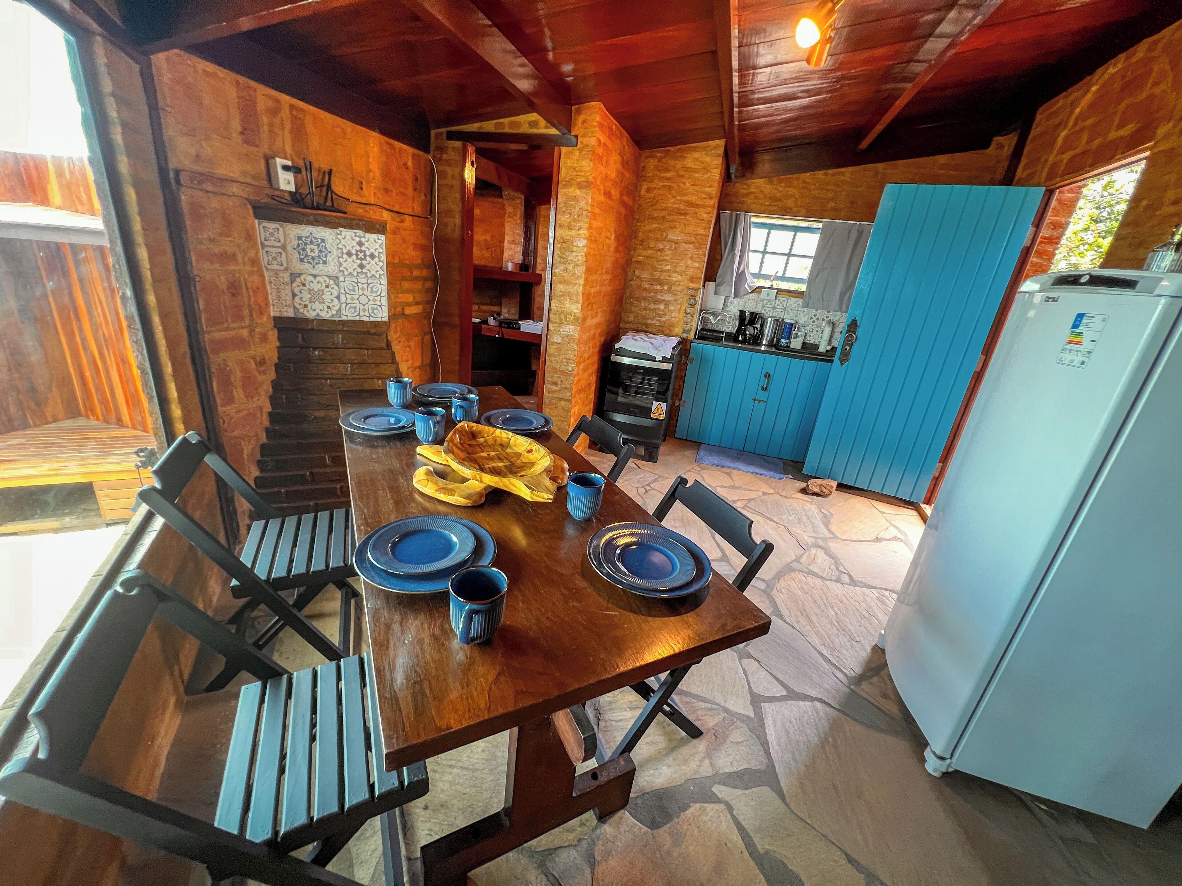 Uma área de jantar rústica com uma mesa de madeira posta para quatro pessoas, cercada por paredes de pedra e um armário azul.