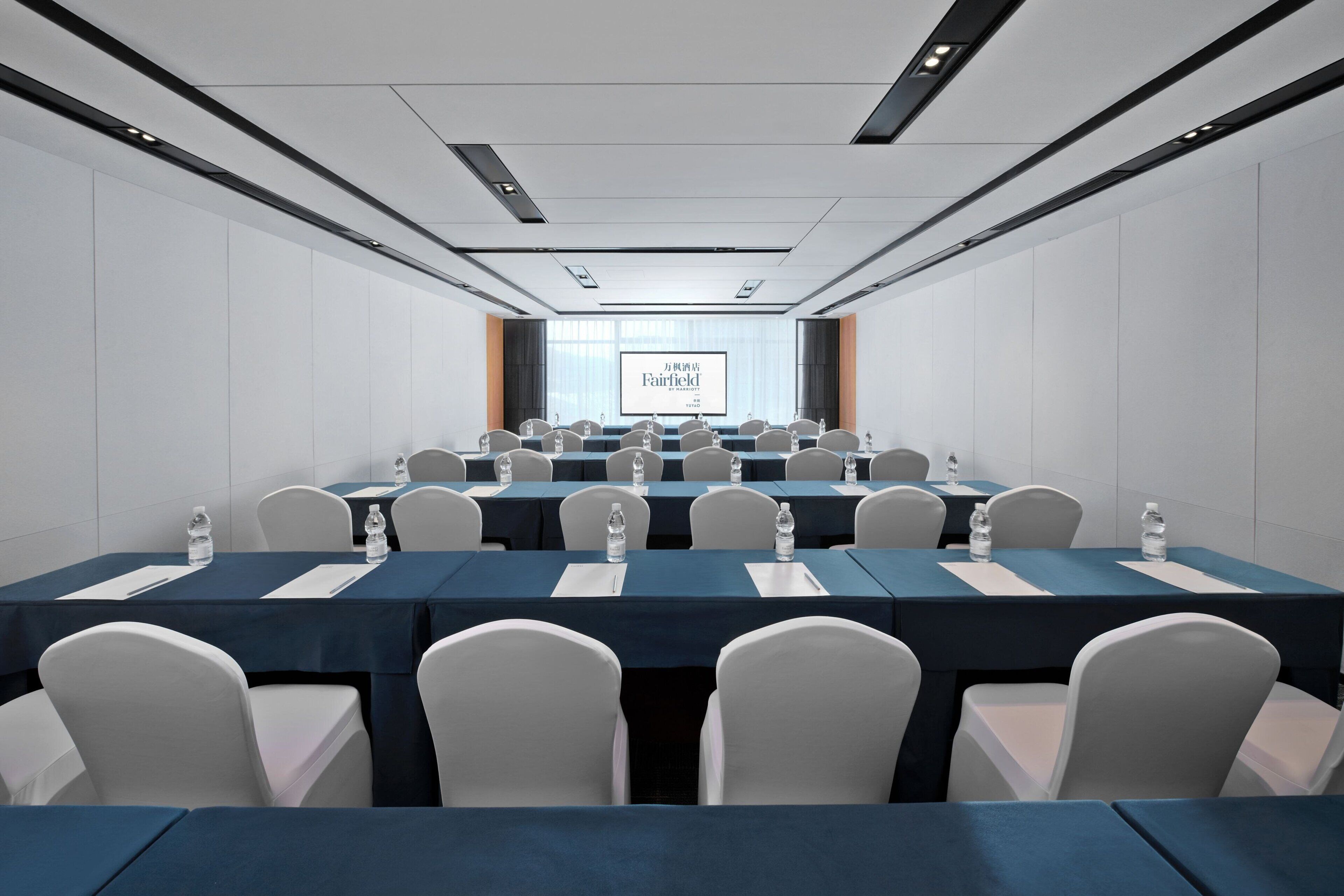 A conference room with multiple tables and chairs, a large screen at the front, and a neutral color scheme.