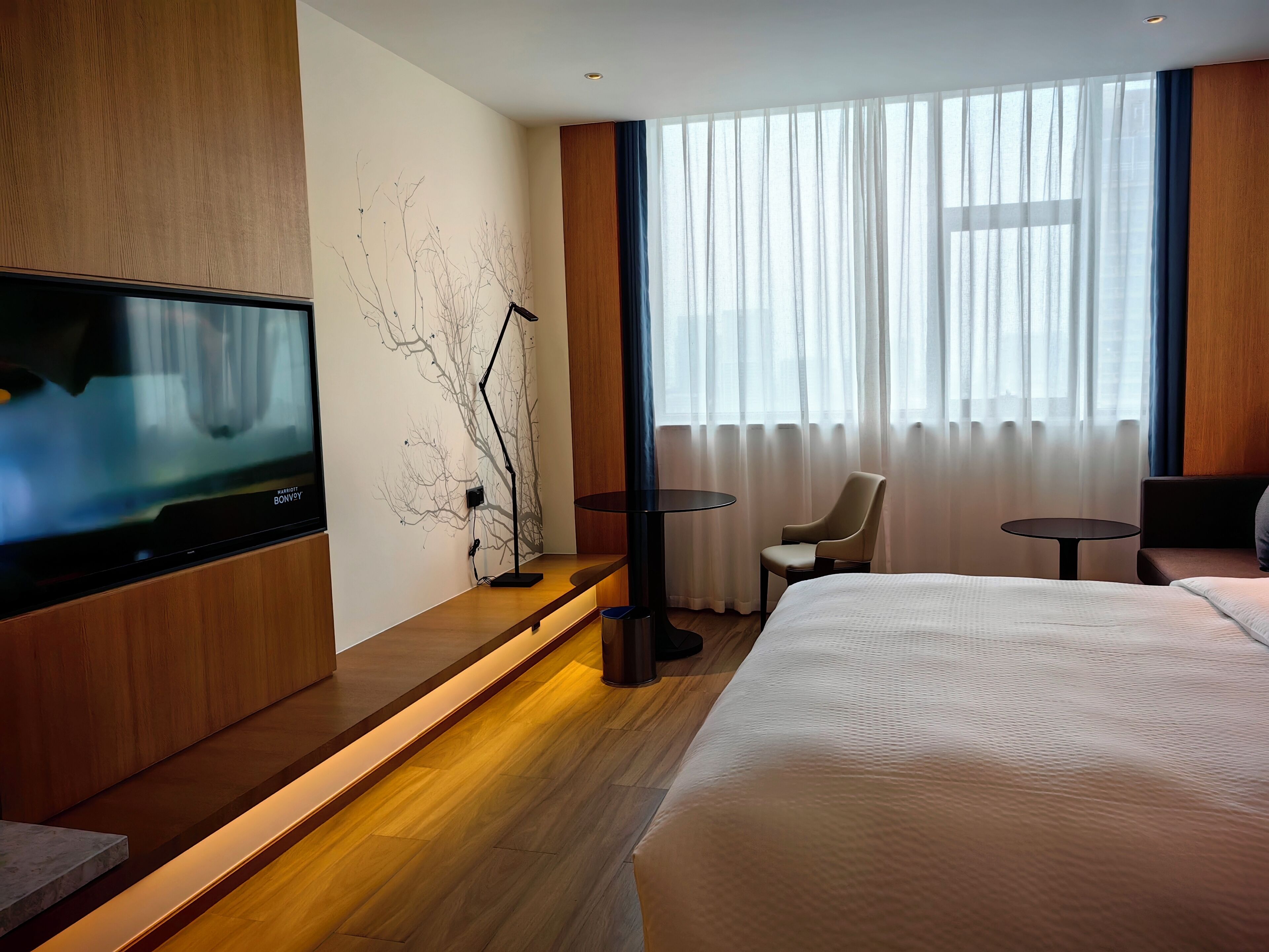 A modern hotel room with a large bed, a flat-screen TV, a small table, and a chair.