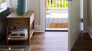 Un plancher en bois avec un paillasson, une lampe bleue posée sur une table d’appoint et une porte donnant sur un balcon.
