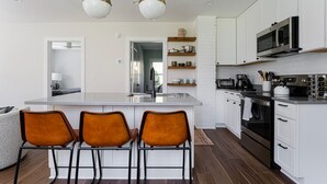Une cuisine moderne avec des meubles blancs, un îlot central doté de tabourets de bar, des appareils en acier inoxydable et un éclairage suspendu.