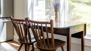 Un coin repas avec une table et des chaises en bois, un vase rempli de fleurs et une vue sur l’extérieur grâce à de grandes fenêtres.
