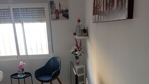 Ett rum med ett soffbord i glas, en blå stol, ett litet bord med en rosa växt och en vägghylla med dekorativa föremål.