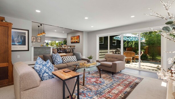 A modern living room with a sofa, armchairs, a coffee table, and a rug. There is a dining area with chairs and a table in the background.