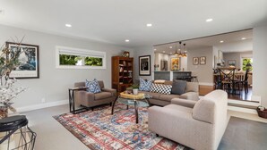 A spacious living room with two sofas, a coffee table, and a rug. There is a bookshelf and a dining area visible in the background.