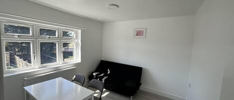 A room with a white table and chairs, a black sofa, a radiator, and a window with a view of trees and a fence.