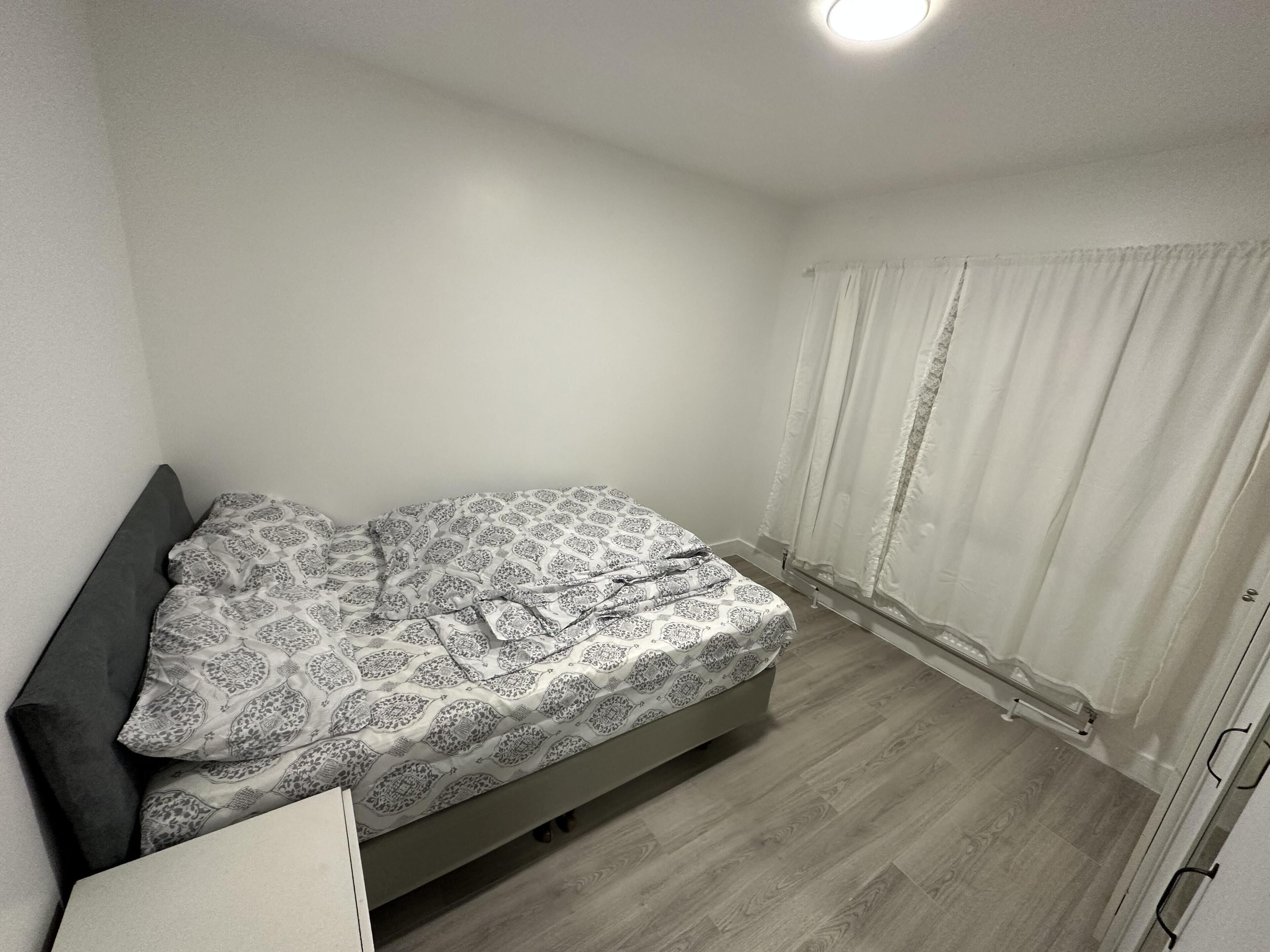 Una habitación pequeña con una cama individual, una colcha estampada, un cabecero oscuro y una ventana con cortinas translúcidas.