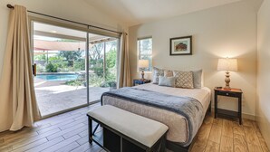 Een slaapkamer met een groot bed, een bankje, een nachtkastje met een lamp, en uitzicht op een zwembad door een schuifdeur.