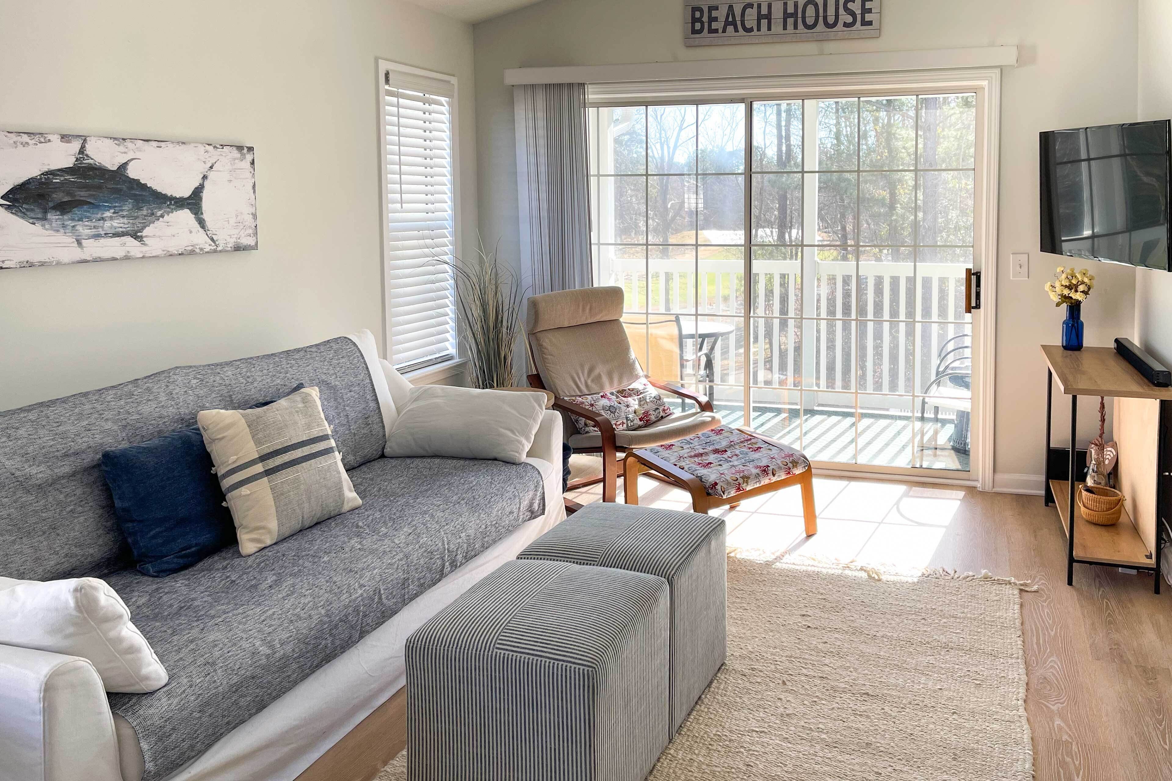A living room with a grey sofa, a striped ottoman, a wooden chair, and a TV on a wooden stand.