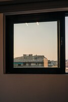Janela com vista para um conjunto de edifícios, incluindo uma estrutura grande com vários andares e um prédio menor com antena parabólica.