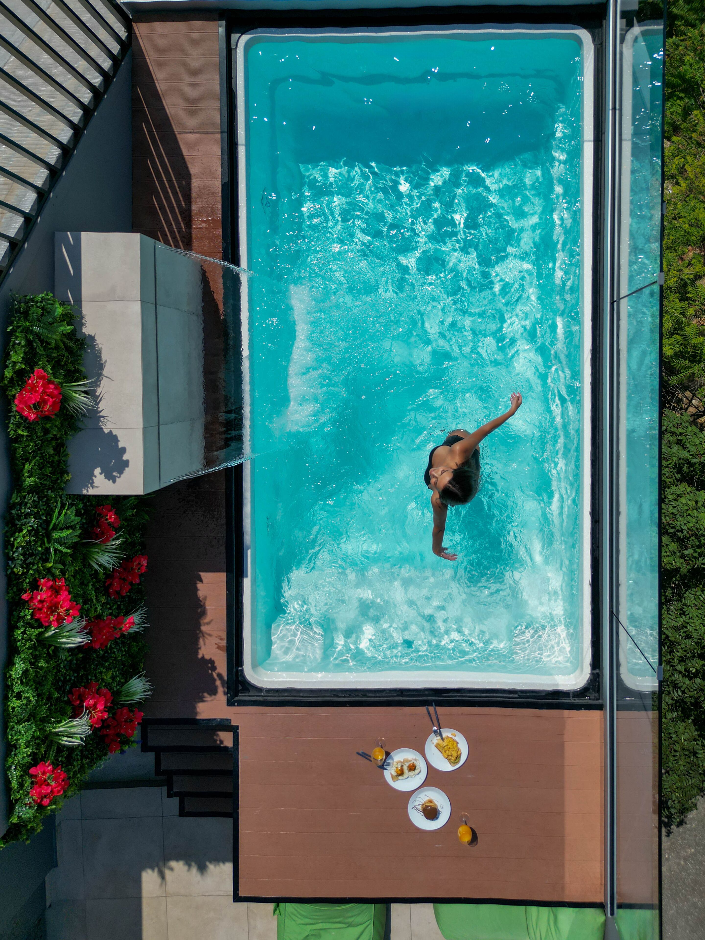 Piscina rectangular con una persona nadando, rodeada por una terraza de madera y flores rojas.