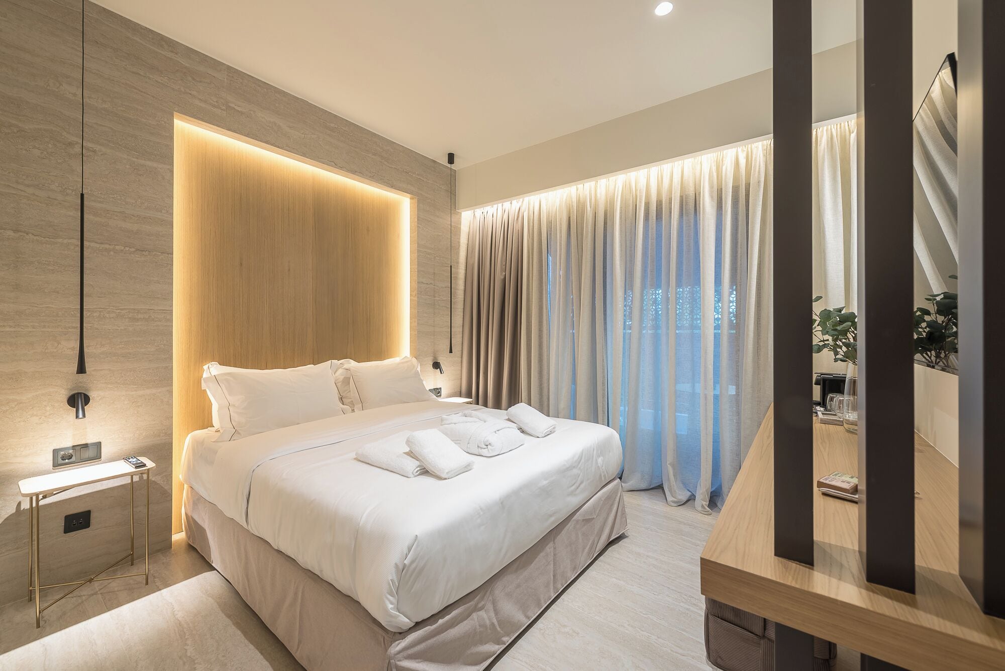 Ein modernes Hotelzimmer mit einem großen Bett, Nachttischen, einer integrierten Lichtleiste und Blick ins Freie.