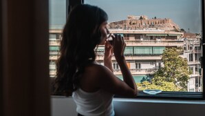 Une personne se tient près d’une fenêtre, d’où l’on peut admirer un bâtiment historique et, au loin, des immeubles modernes.