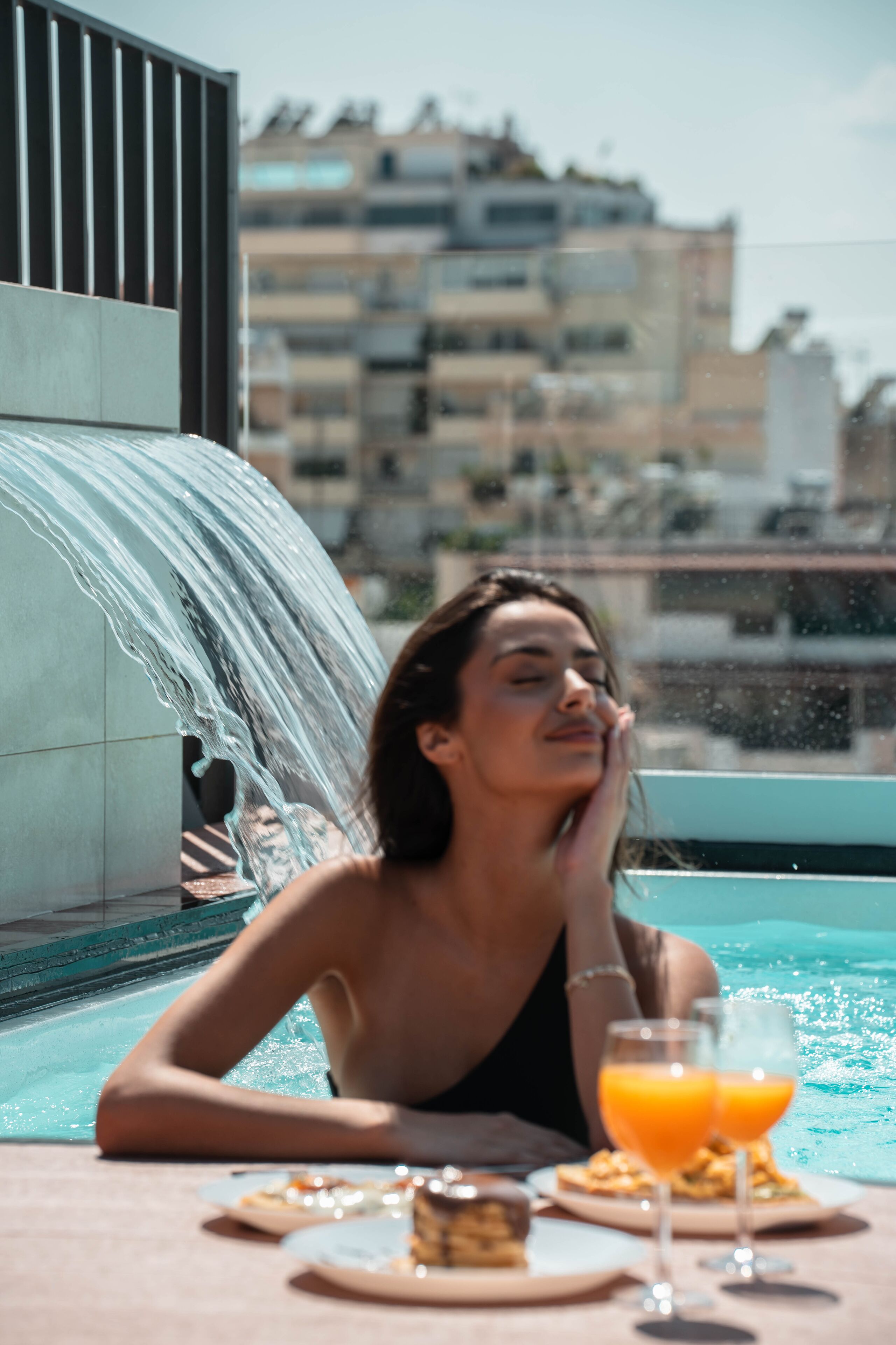A woman relaxing by a pool with a waterfall feature, dining table with plates and drinks, and a cityscape in the background.