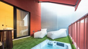 Un área moderna para jacuzzi al aire libre con césped artificial, dos almohadas blancas y un toldo rojo.