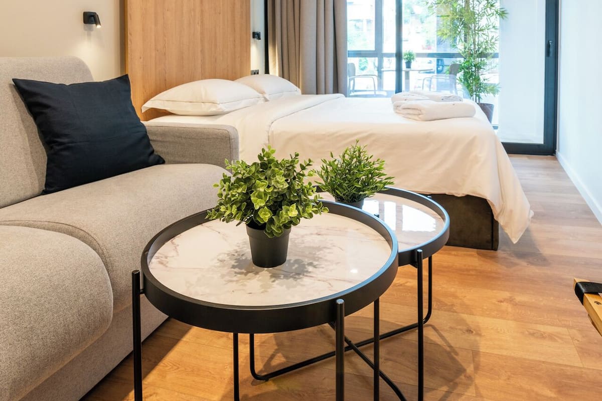 Una habitación de hotel moderna con sofá, dos camas, una mesa redonda con plantas y un ventanal con vistas a la ciudad.
