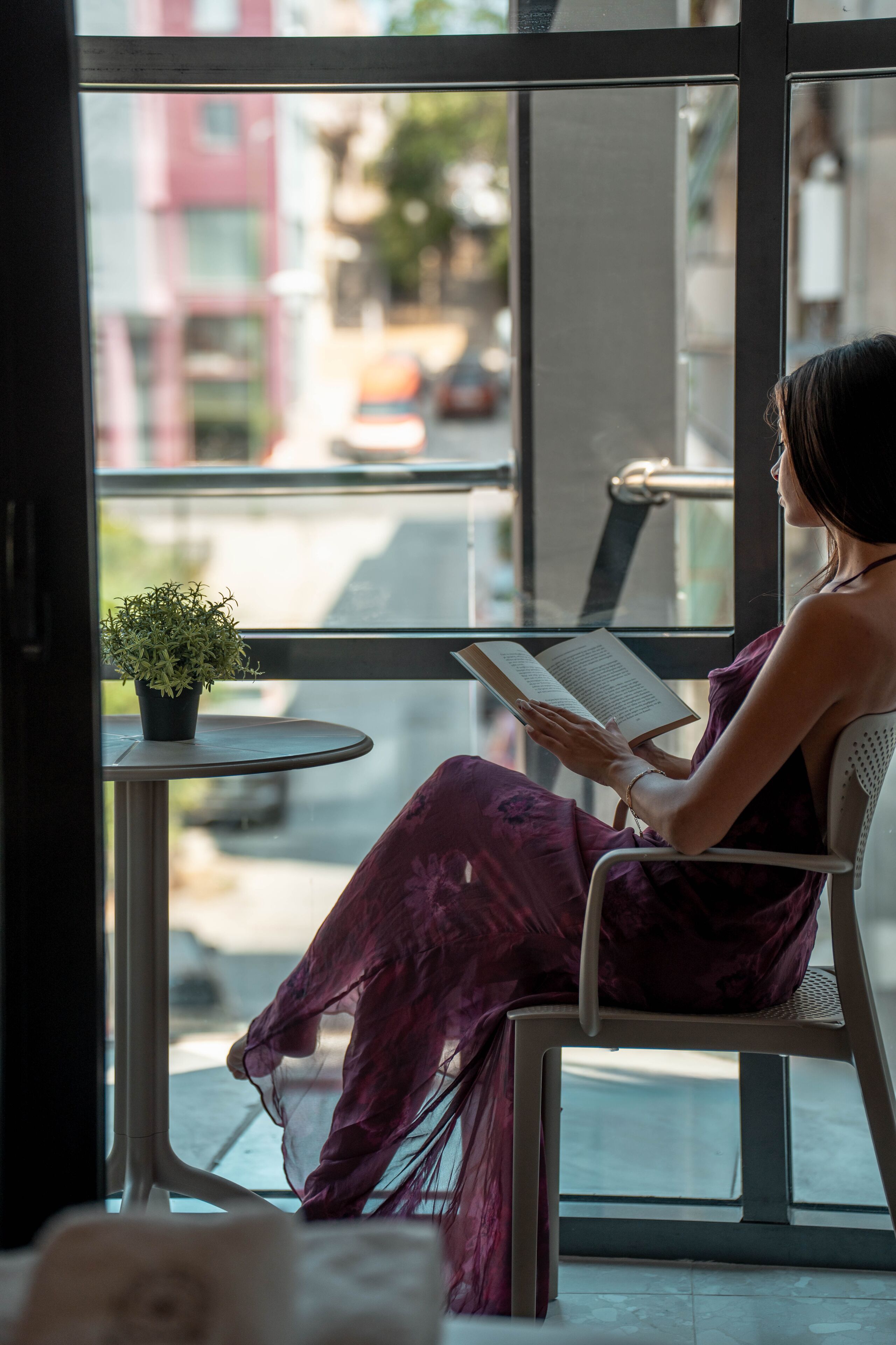 Une personne assise près d’une fenêtre, en train de lire un livre, avec une petite table et une plante en pot à proximité.