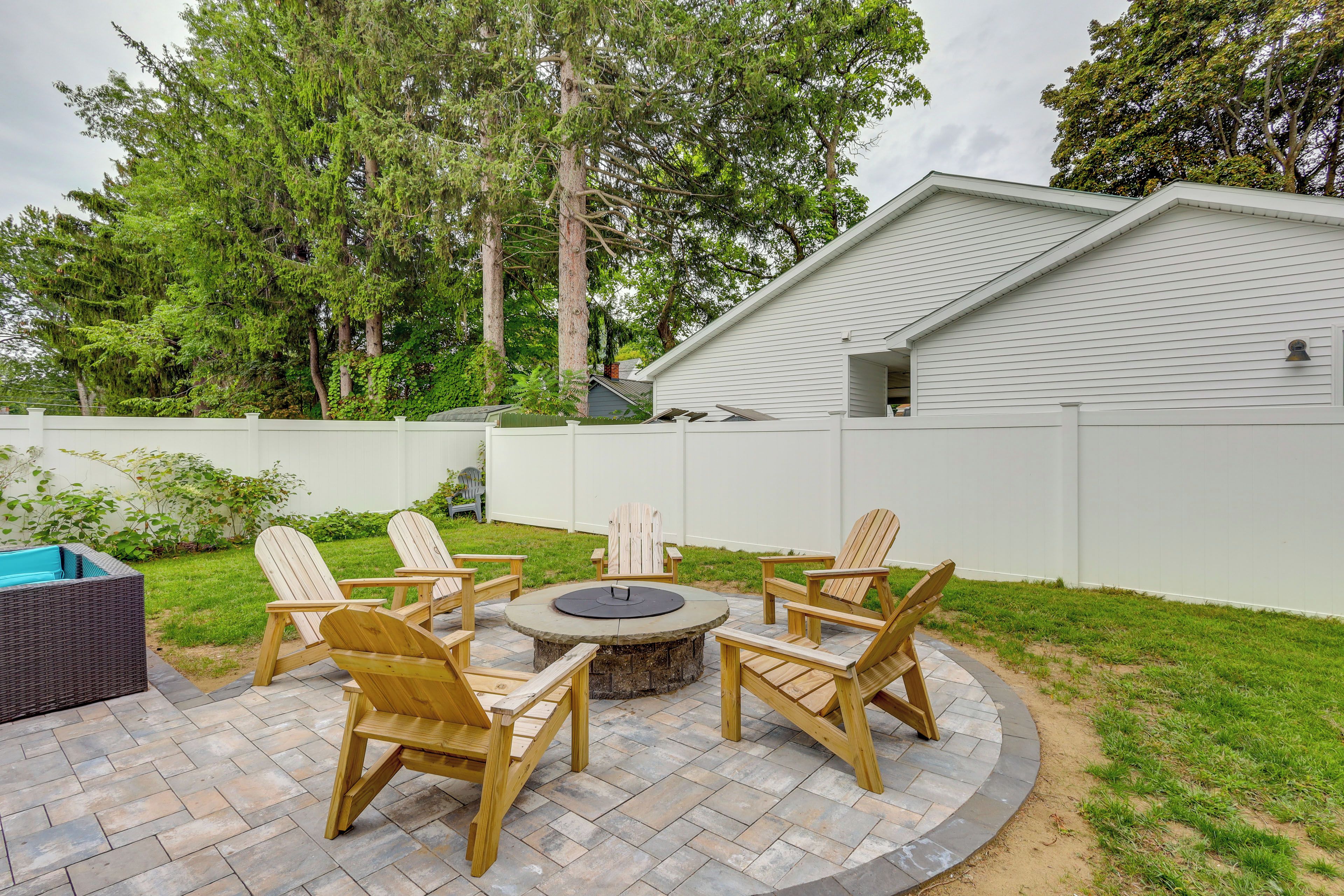 A backyard with a circular fire pit table surrounded by wooden Adirondack chairs.
