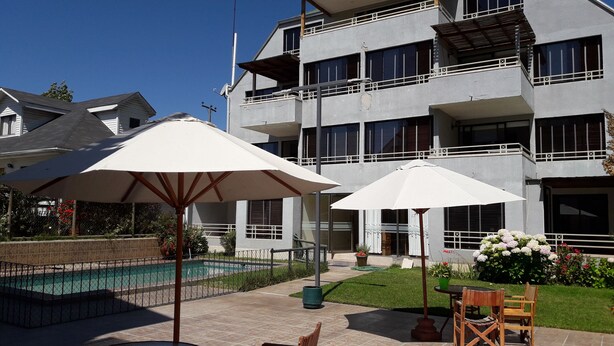 A poolside area with outdoor seating and umbrellas, a swimming pool, and a multi-story building in the background. - Habitamar Maitencillo (Puchuncavi)